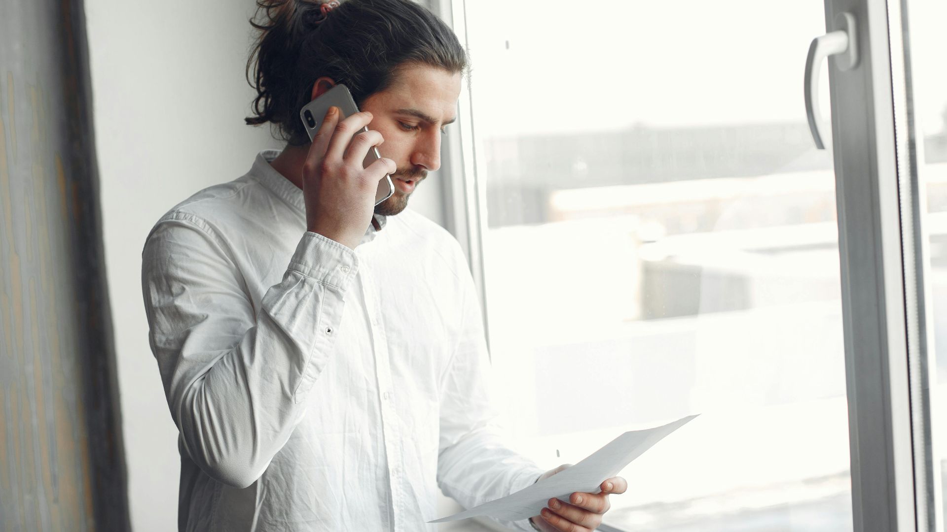 Young man in white shirt, on phone call holding a document, standing by a large window.