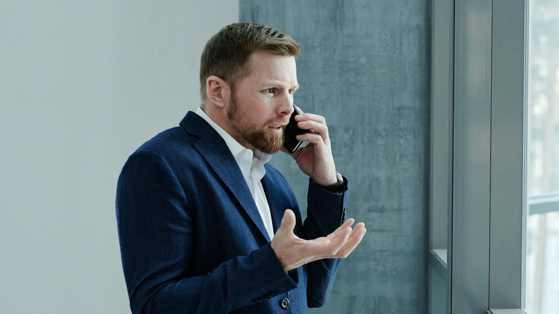 Businessman using smartphone while standing indoors by glass window, expressing concern.