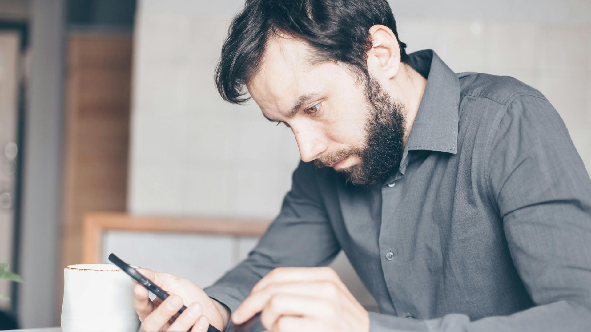 A bearded man intensely stares at his smartphone, deep in thought, indoors.