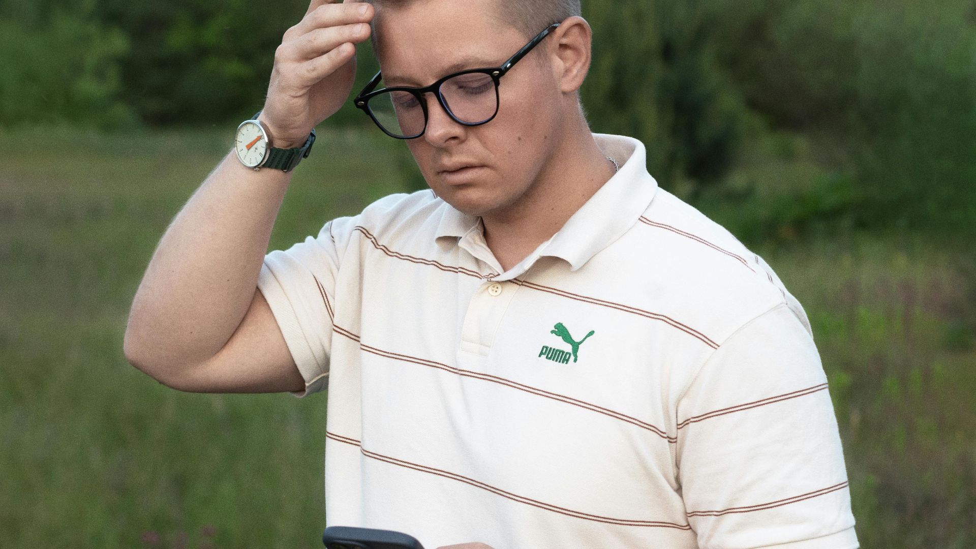 A young man in casual wear checking his smartphone in a natural outdoor setting, looking thoughtful.