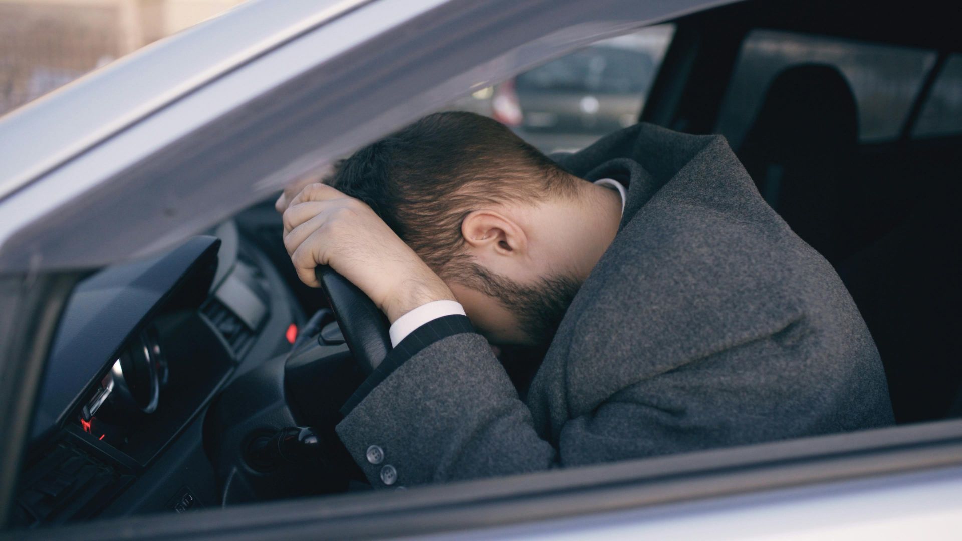 Man feeling overwhelmed and stressed, resting his head on a steering wheel inside a car.