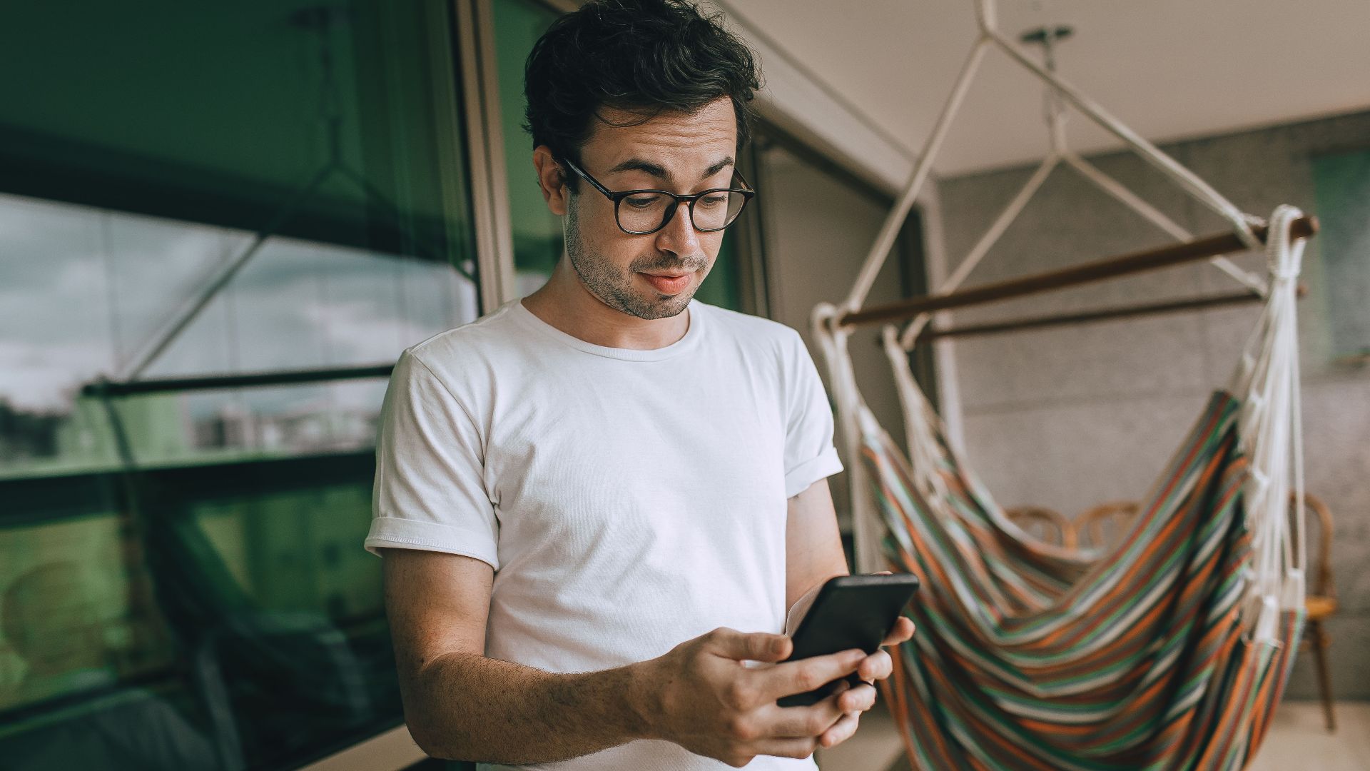 Young man in glasses using smartphone indoors with striped hammock.
