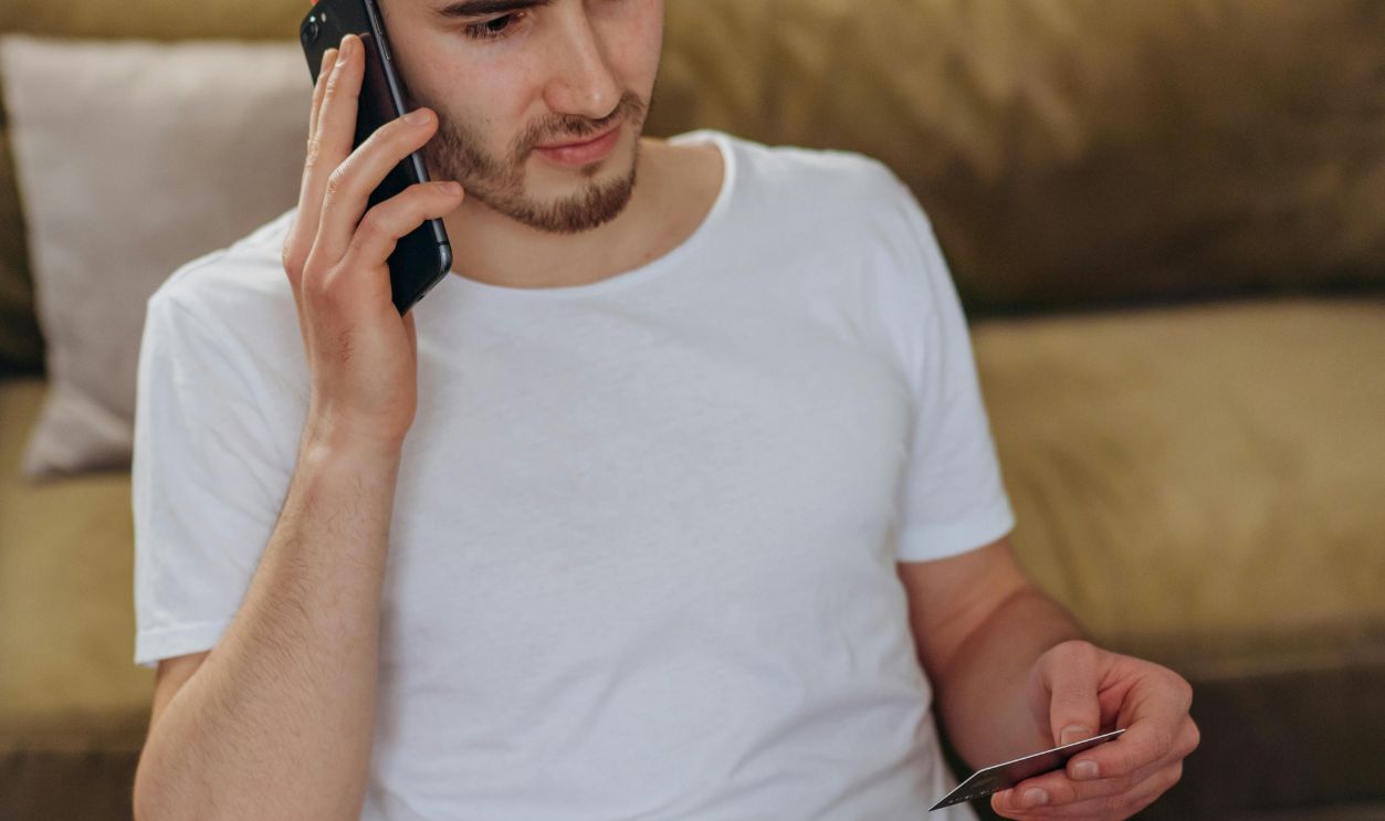 Young man using phone and credit card for online shopping, seated indoors.