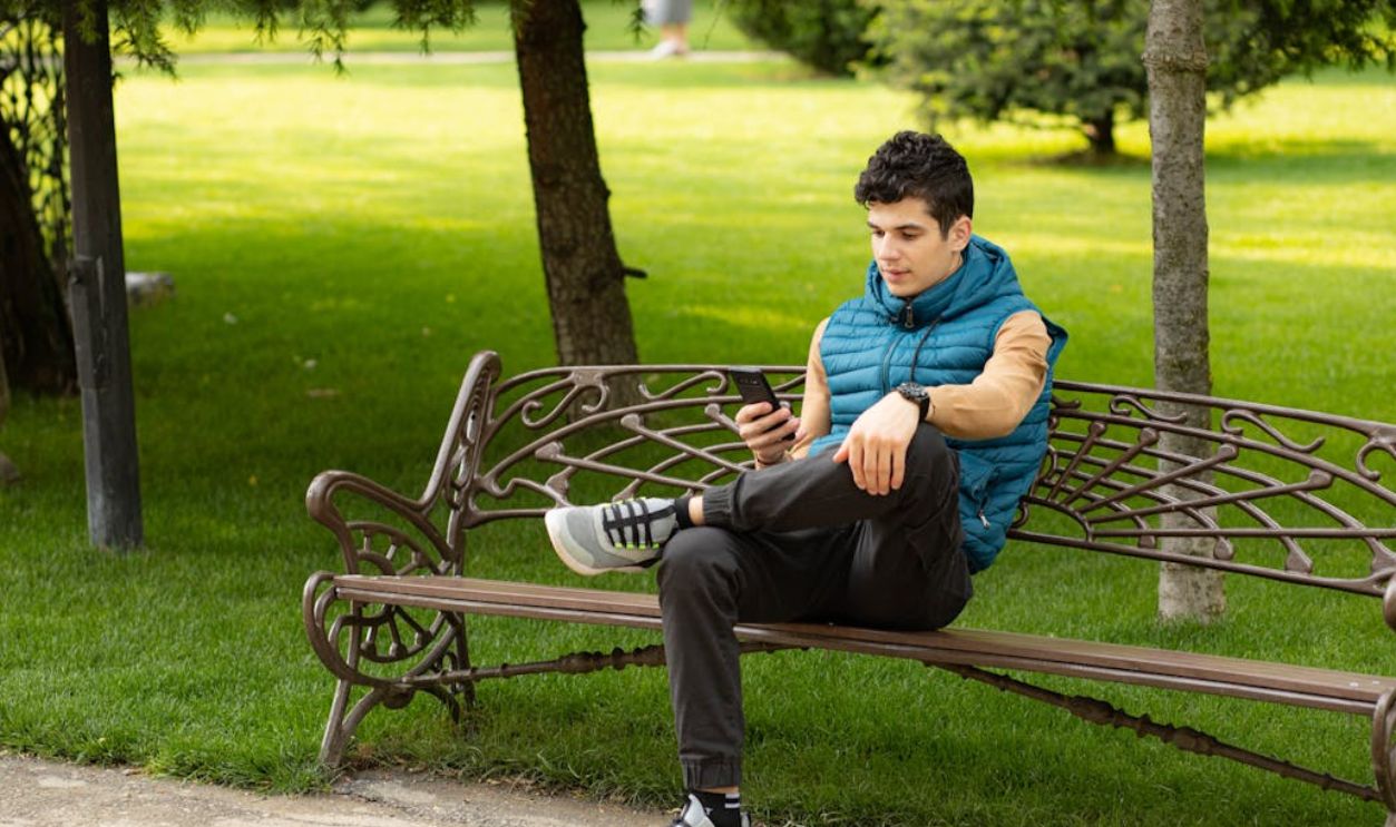 Teenage boy in a blue jacket looks at his smartphone while sitting on a park bench in a sunlit garden