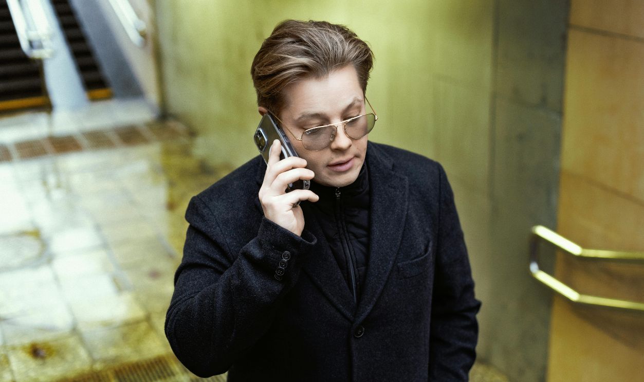 A man in a coat speaks on his smartphone as he walks through an urban environment.