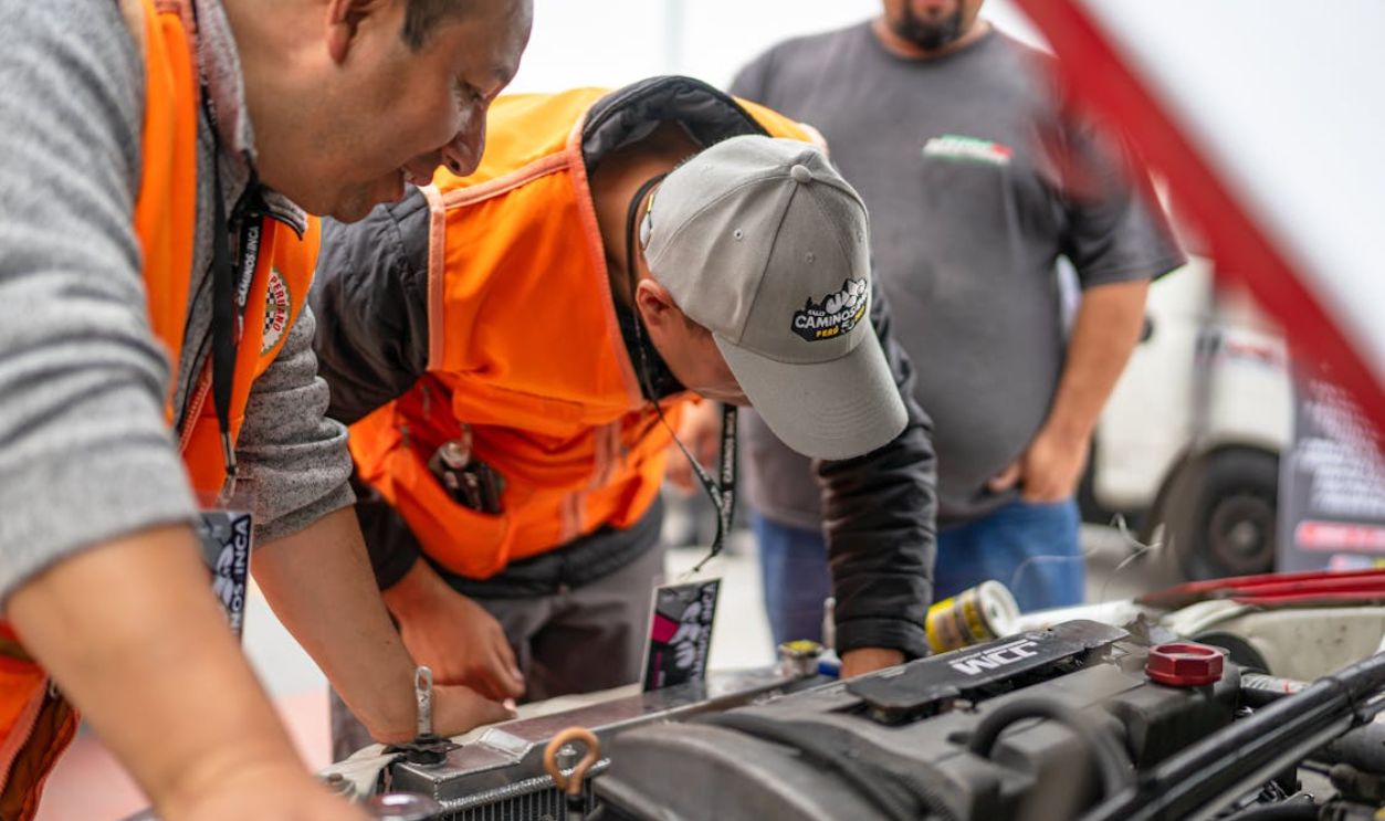 Mechanics focused on inspecting vehicle engine with open hood, wearing safety vests.