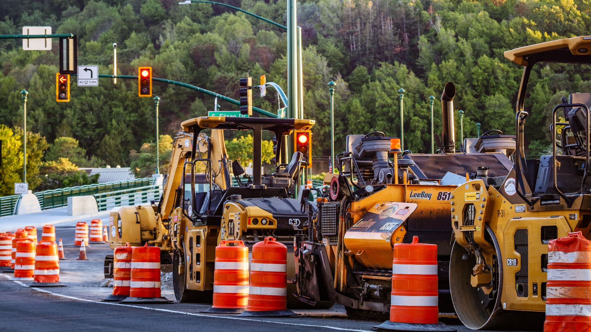 Heavy machinery and barriers at a road construction site with traffic lights and forest backdrop.