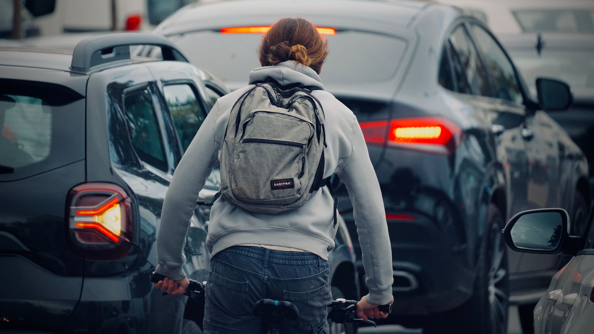 A cyclist wearing a backpack rides through a congested city traffic jam during daytime.