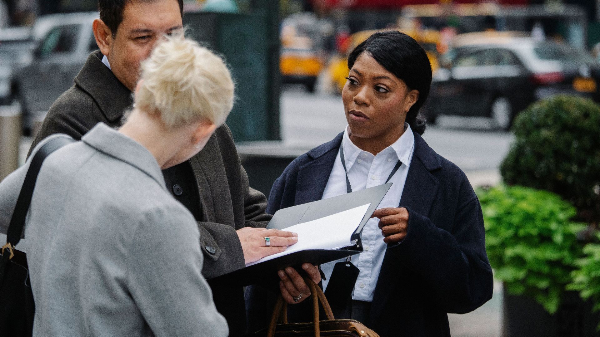 Focused multiracial colleagues in elegant formal wear standing with opened paper folder and discussing business strategy on urban street
