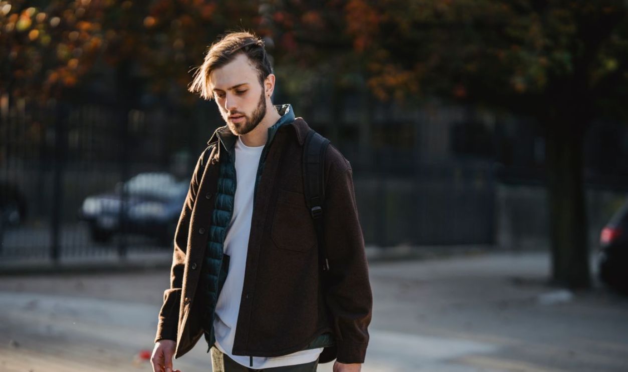 A young man walks through a sunlit park in fall, wearing casual autumn attire and a backpack.