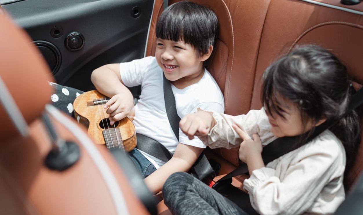 Two happy children in a convertible car playing with a ukulele during a sunny trip.