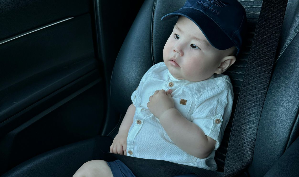 Cute toddler wearing a cap and shoes, seated safely in a car seat, looking curious.