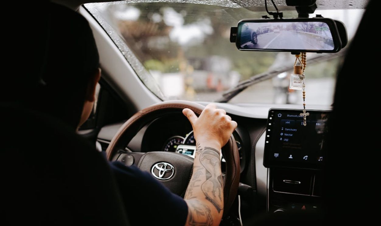 A driver's tattooed arm on the steering wheel inside a Toyota car during the day.