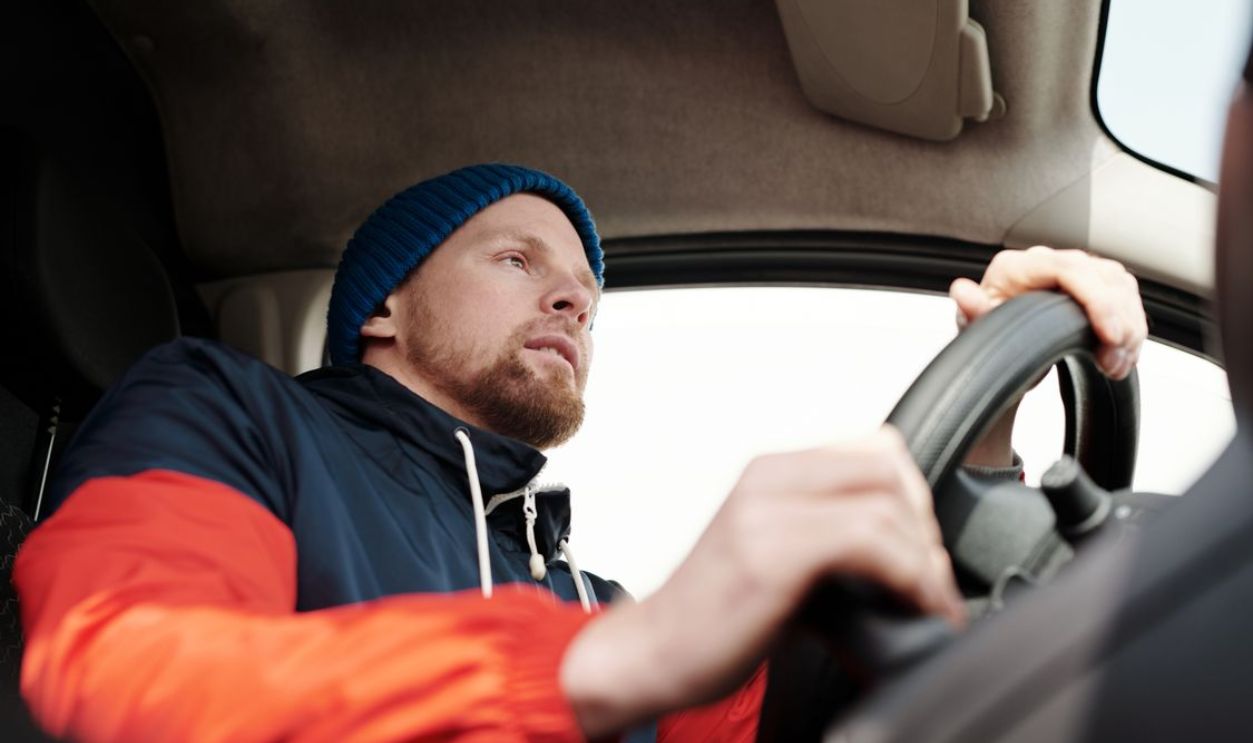 Low angle shot of a bearded man driving a car, wearing a hoodie and knit cap.