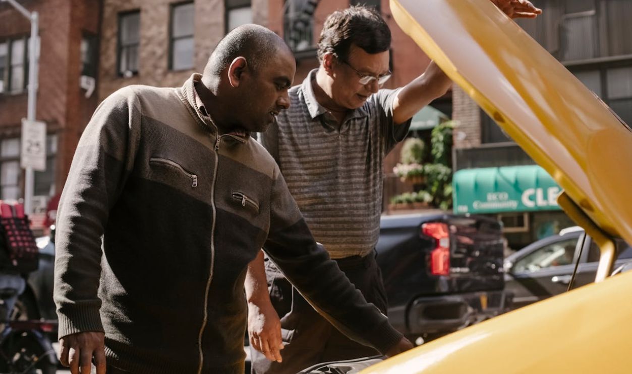 Two men discussing a car engine issue in a city street setting.