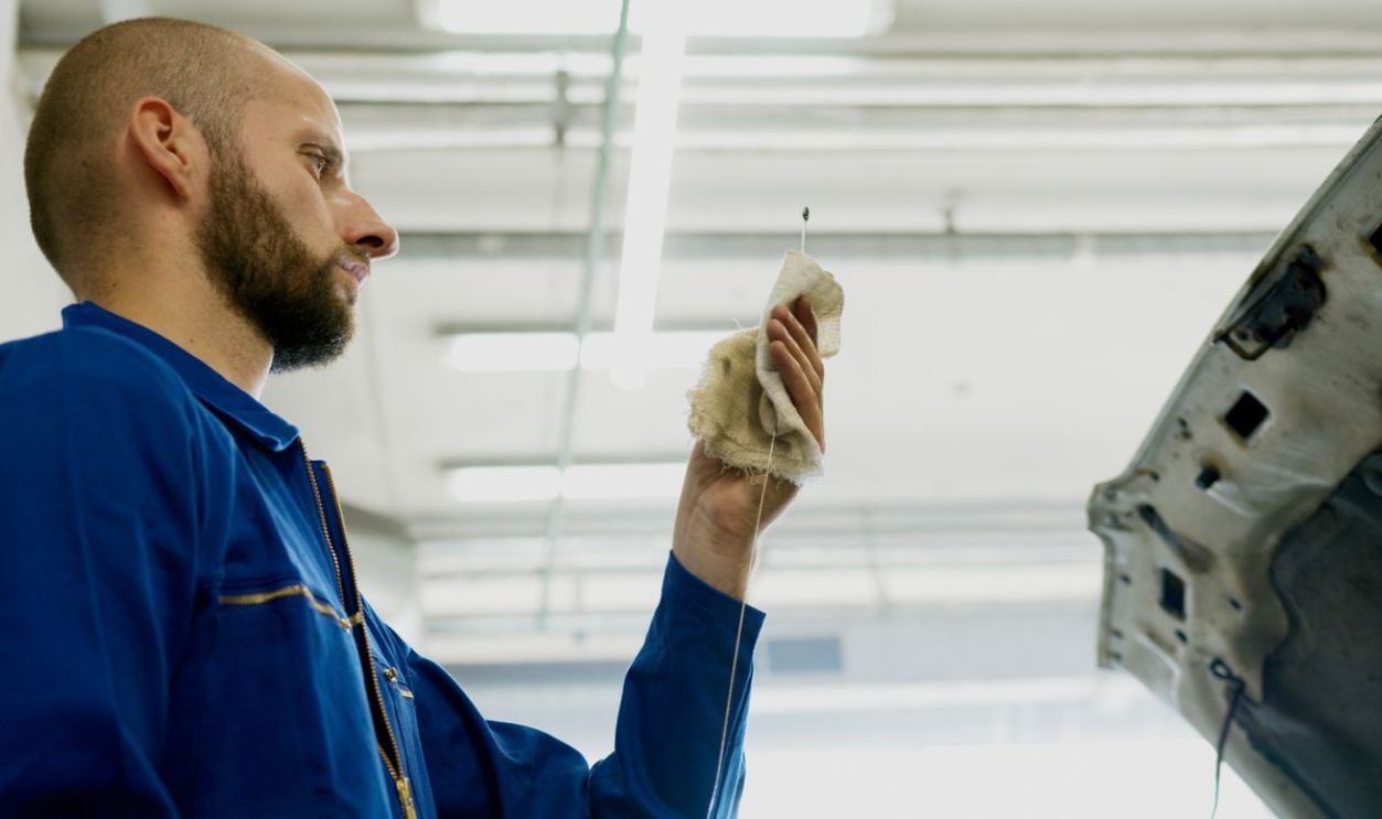 Mechanic in a blue uniform checks car oil level using a dipstick inside a garage.