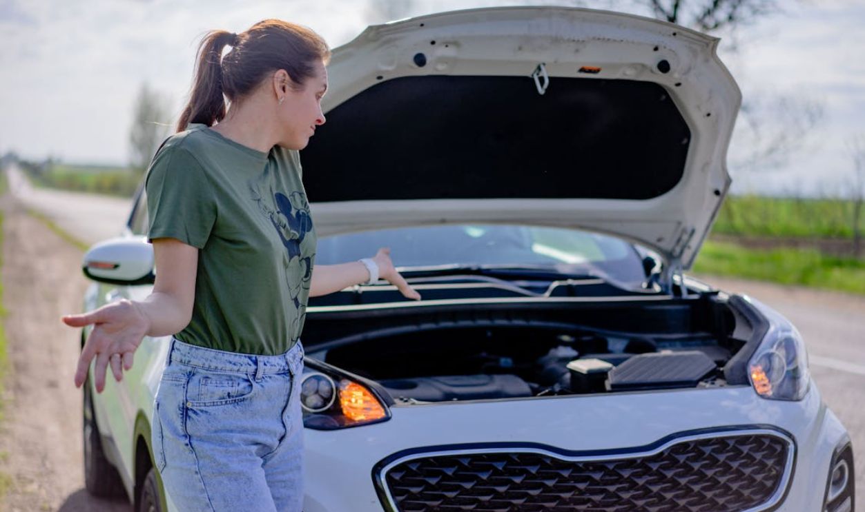 A woman looks puzzled by her broken car with an open hood on a rural road.