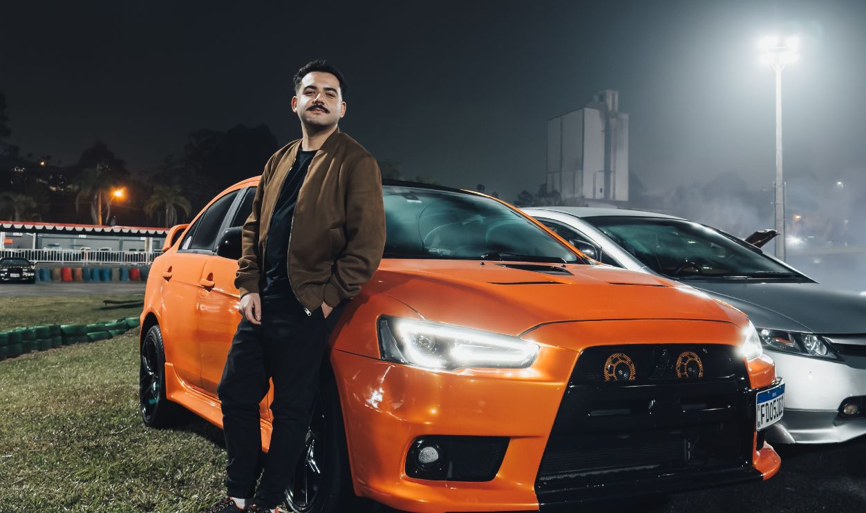 A man stands confidently beside an orange sports car during a nighttime car event, exuding style and enthusiasm