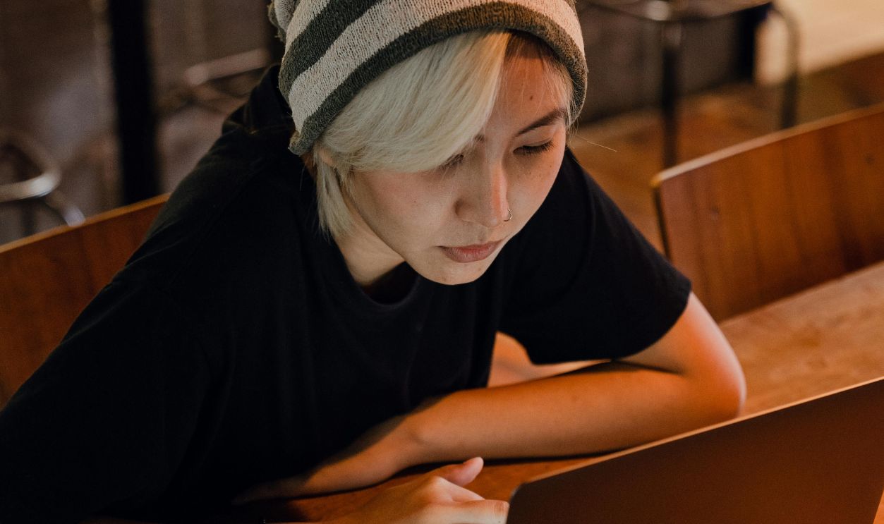 A young woman with a beanie focuses intently on her laptop in a warm, dimly lit cafe setting