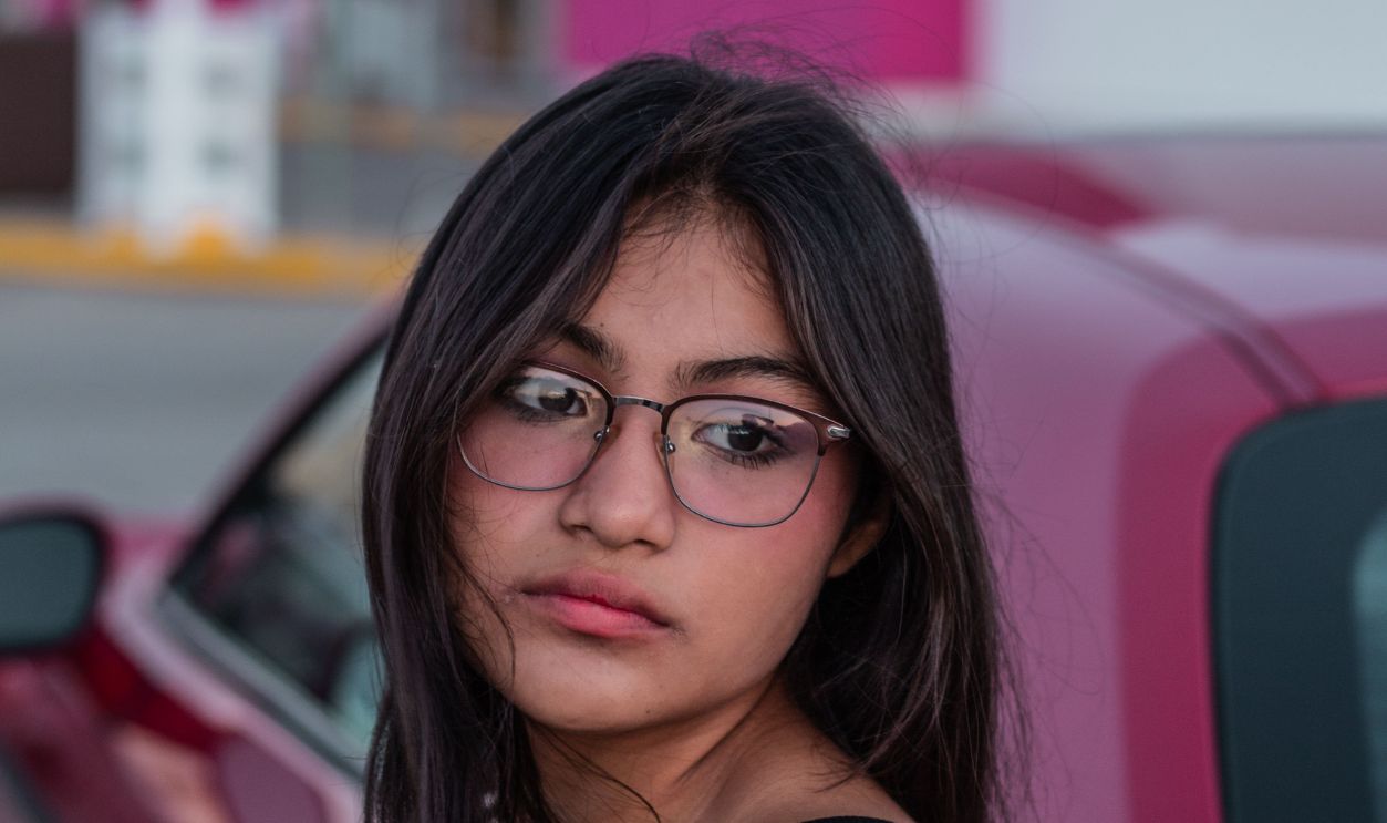 Portrait of a young woman with eyeglasses and black top leaning on a red car. Urban setting