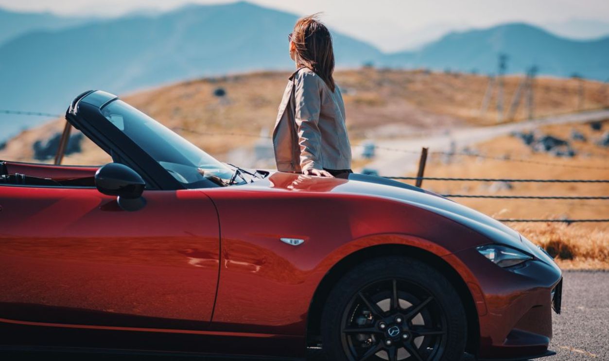 A woman standing by a red convertible sports car, admiring a scenic mountain view