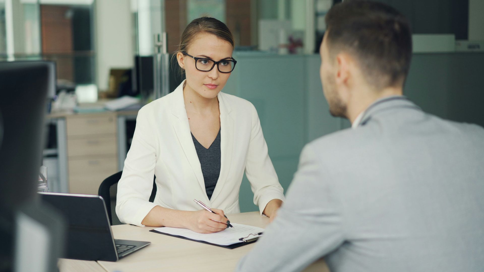 Woman in glasses interviews man at office desk.