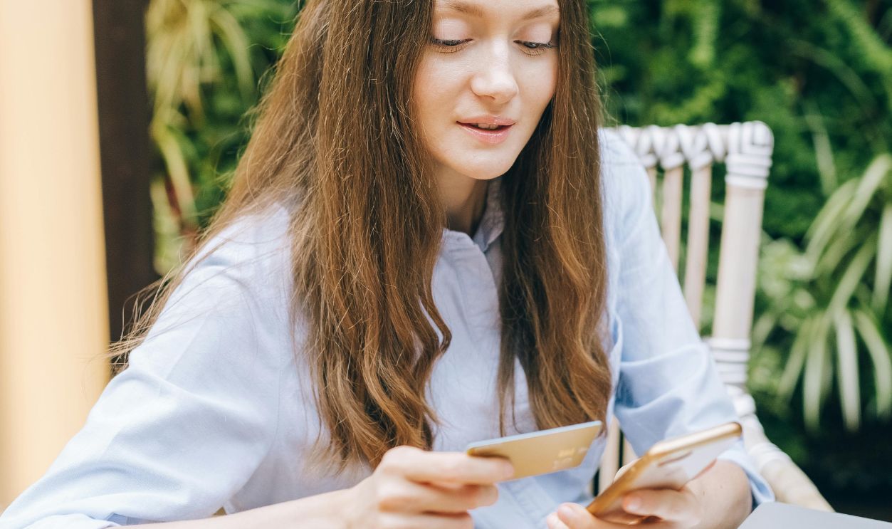 Young woman using smartphone and card for online shopping, sitting at wooden table with laptop
