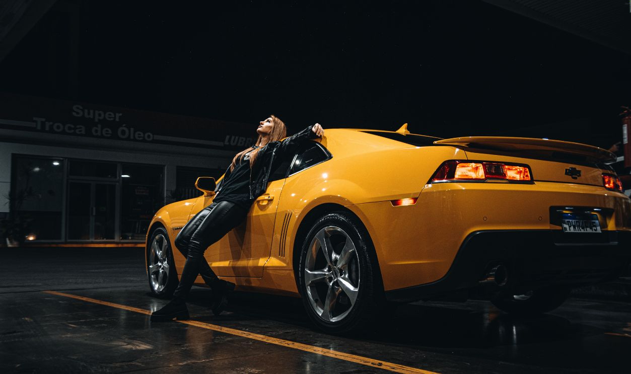 Fashionable woman in leather outfit leaning on a yellow sports car under a starry night sky at a gas station