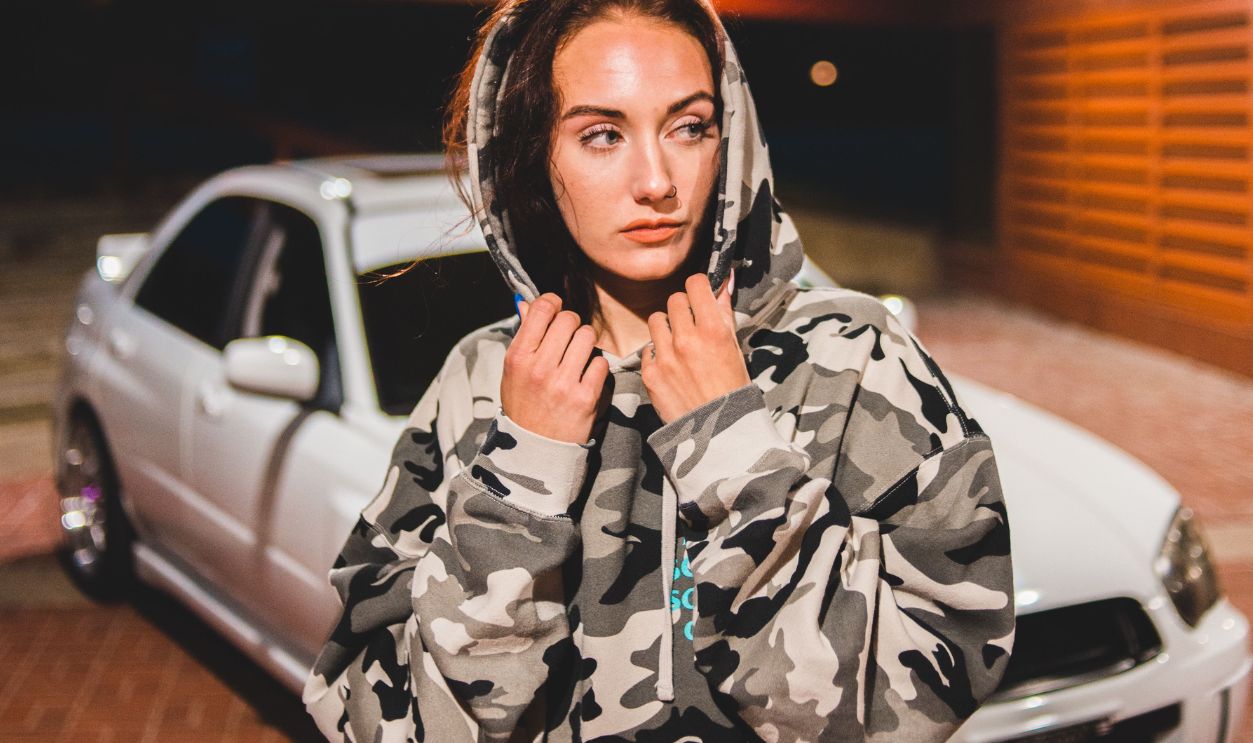 Woman in camo hoodie stands by a white sports car in a dimly lit parking area at night