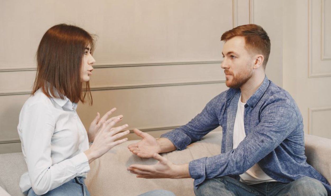 A young couple sitting on a couch having a thoughtful conversation with expressive hand gestures