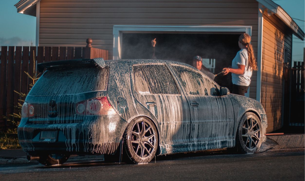A family washes a car with soap suds on a suburban driveway