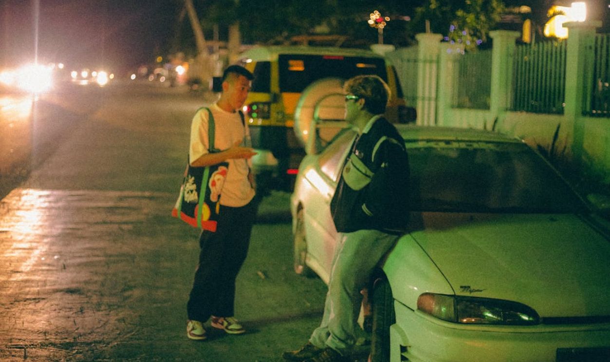 Two young men talking near a car on a dimly lit urban street at night.