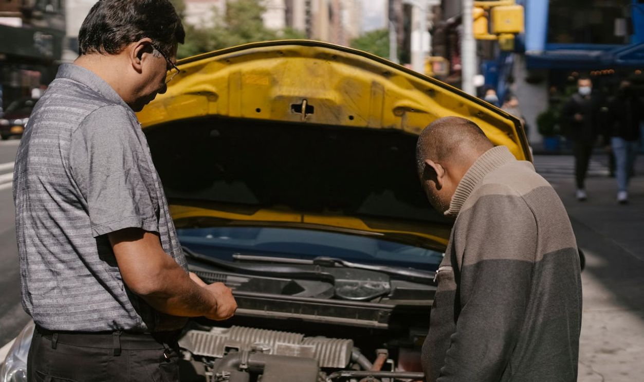 Two men inspecting a car engine on a city street, focusing on vehicle maintenance and repair.