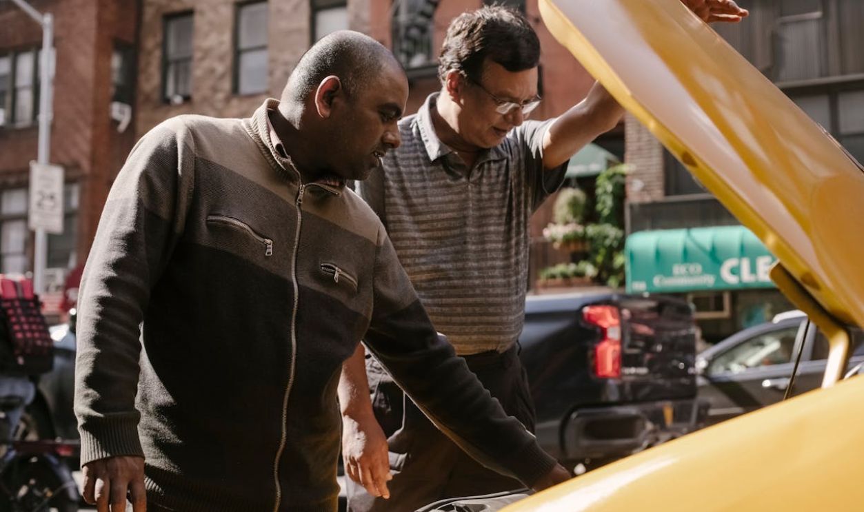 Two men discussing a car engine issue in a city street setting