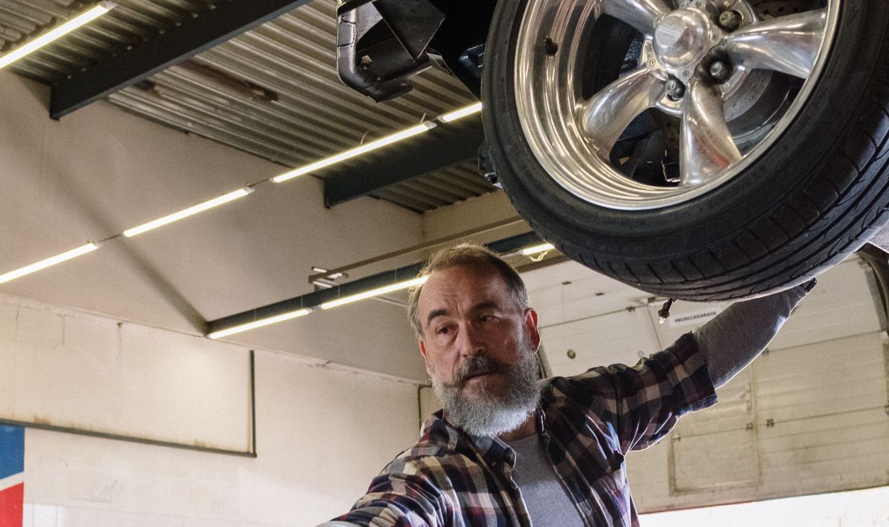 Bearded mechanic working on a vehicle in an auto repair shop, handing a tool to an assistant