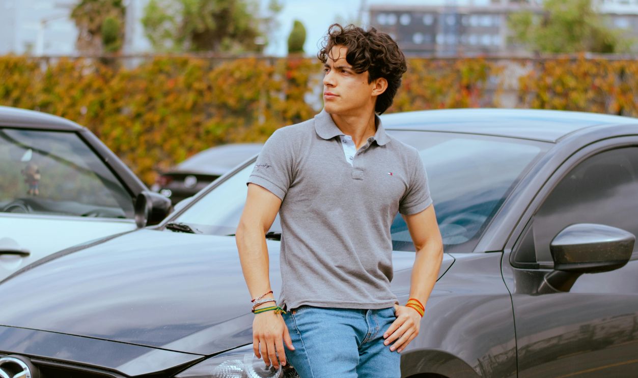 Casual young man posing against a gray car in a city parking area. Modern urban lifestyle