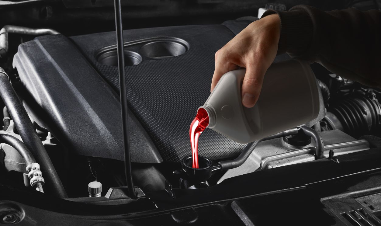 Mechanic hand pouring red transmission fluid into car engine under hood close up. Concept of auto repair, lubricant technology, and professional service visuals for technical communication.