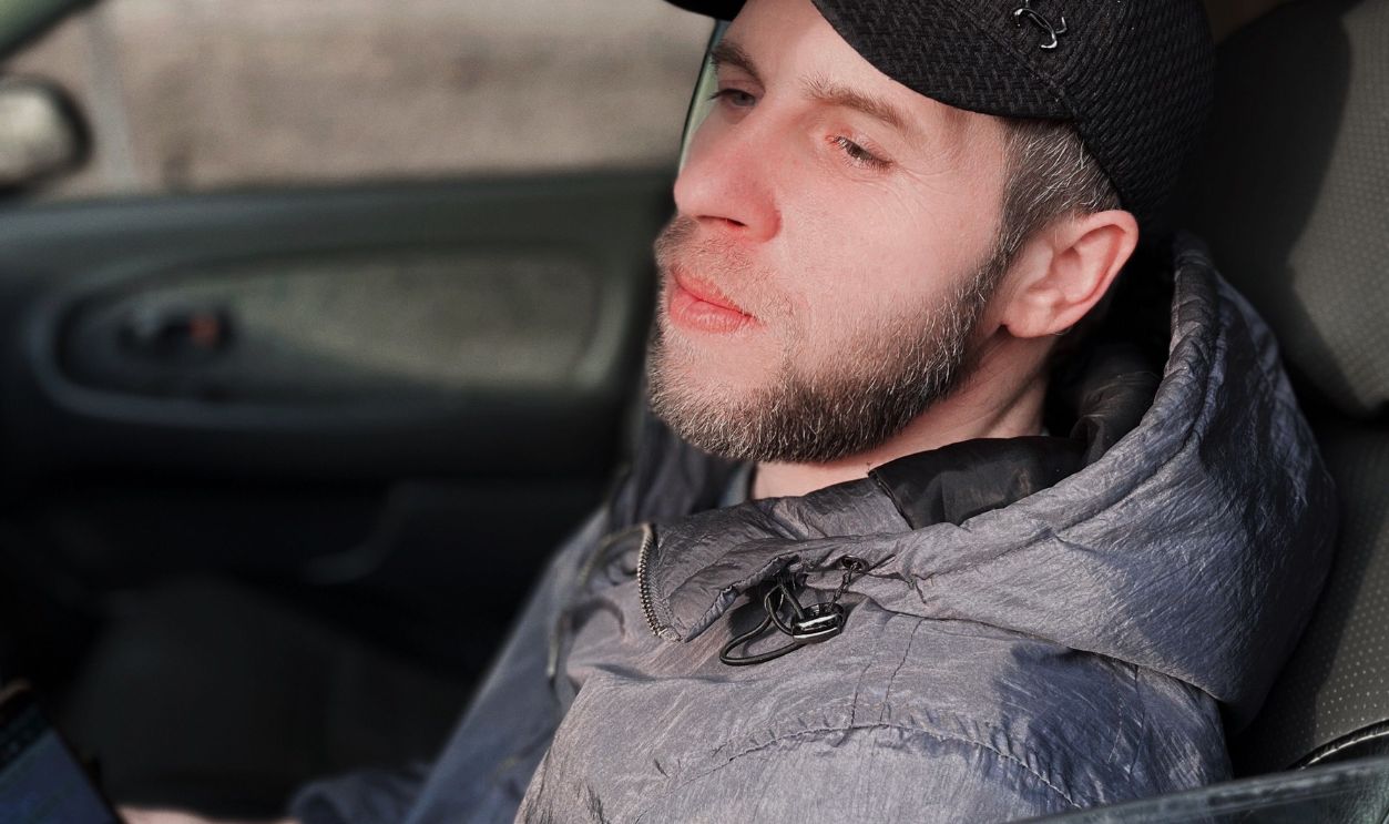 Young man with a beard sitting in a car, looking out while holding a cellphone