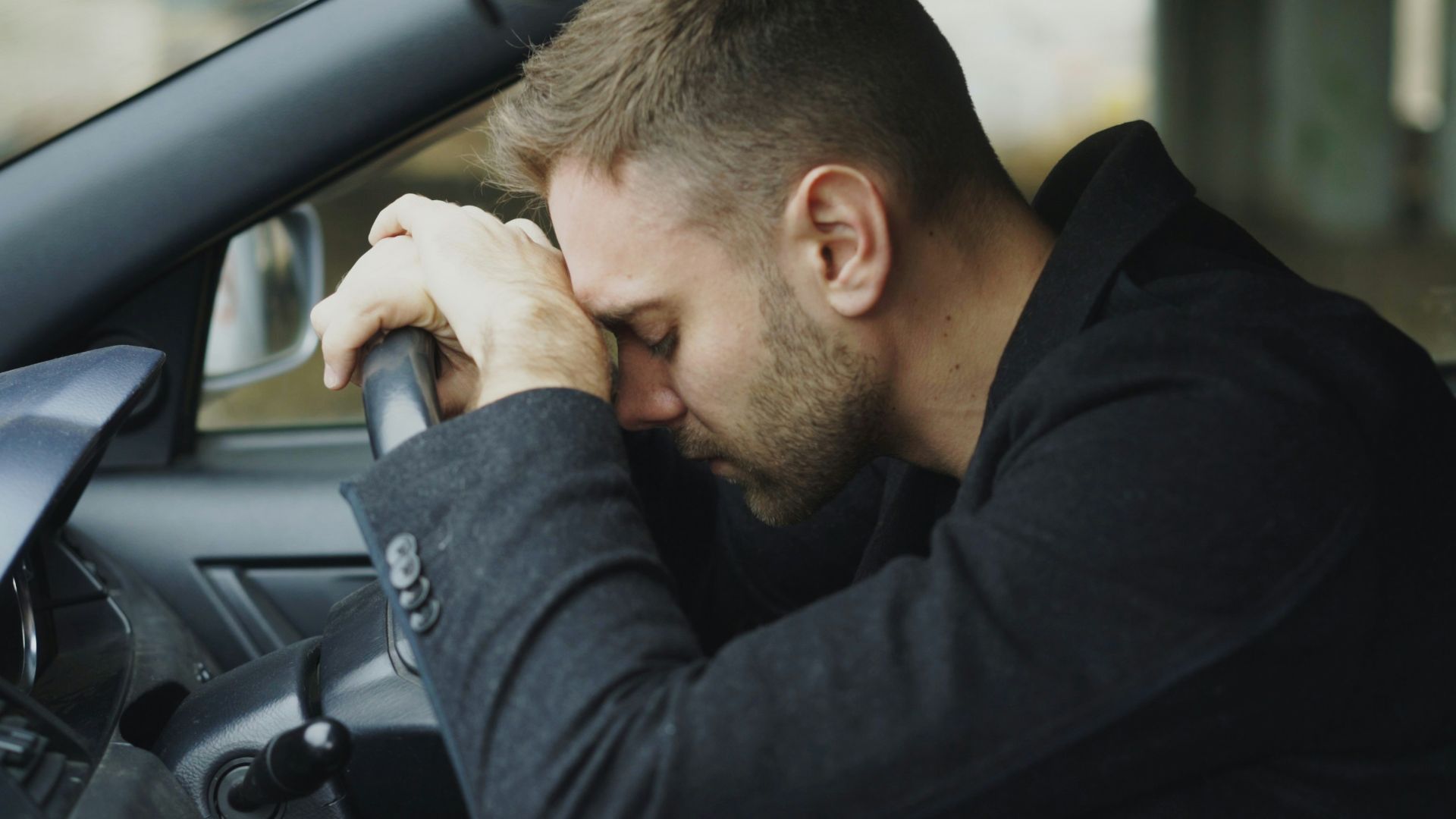 Man resting head on steering wheel in car.