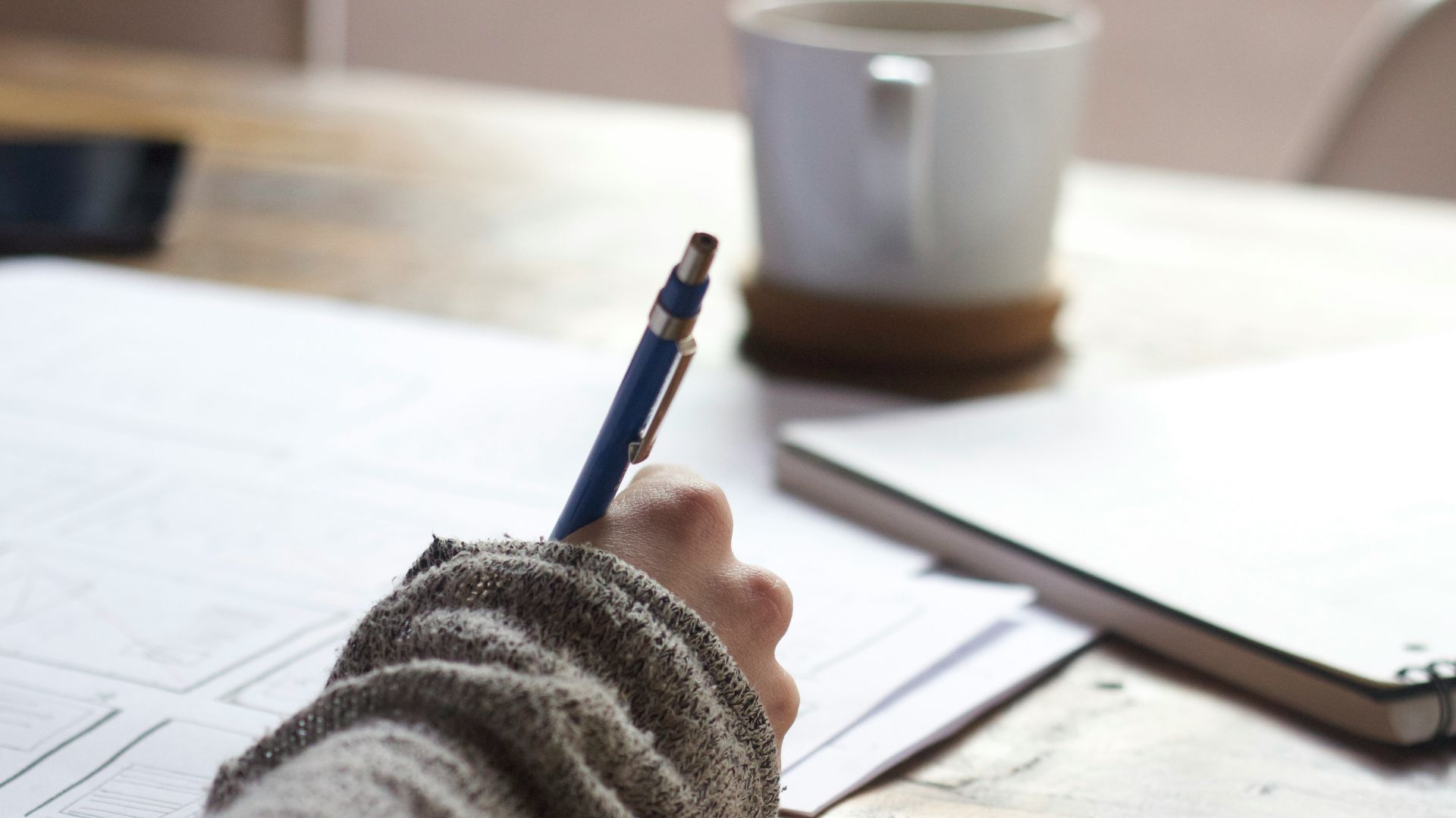 person writing on brown wooden table near white ceramic mug