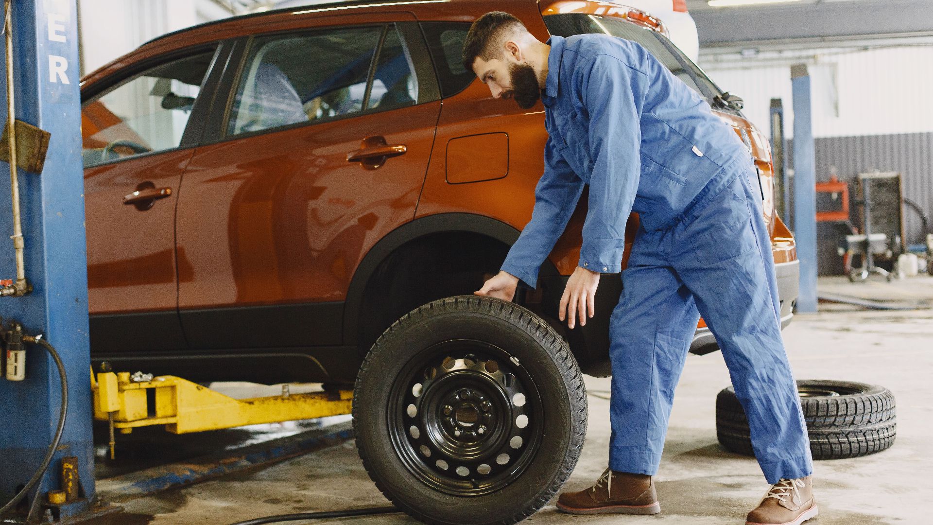 Mechanic in blue coveralls replacing tire on red SUV in repair shop garage.