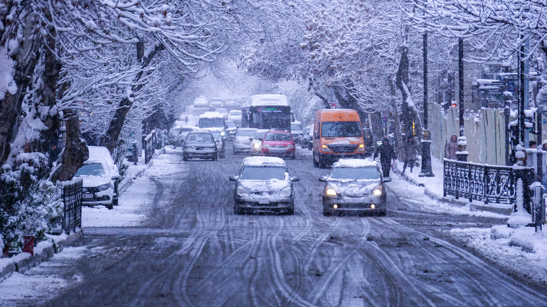 Heavy traffic on a snow-covered urban street lined with bare winter trees.