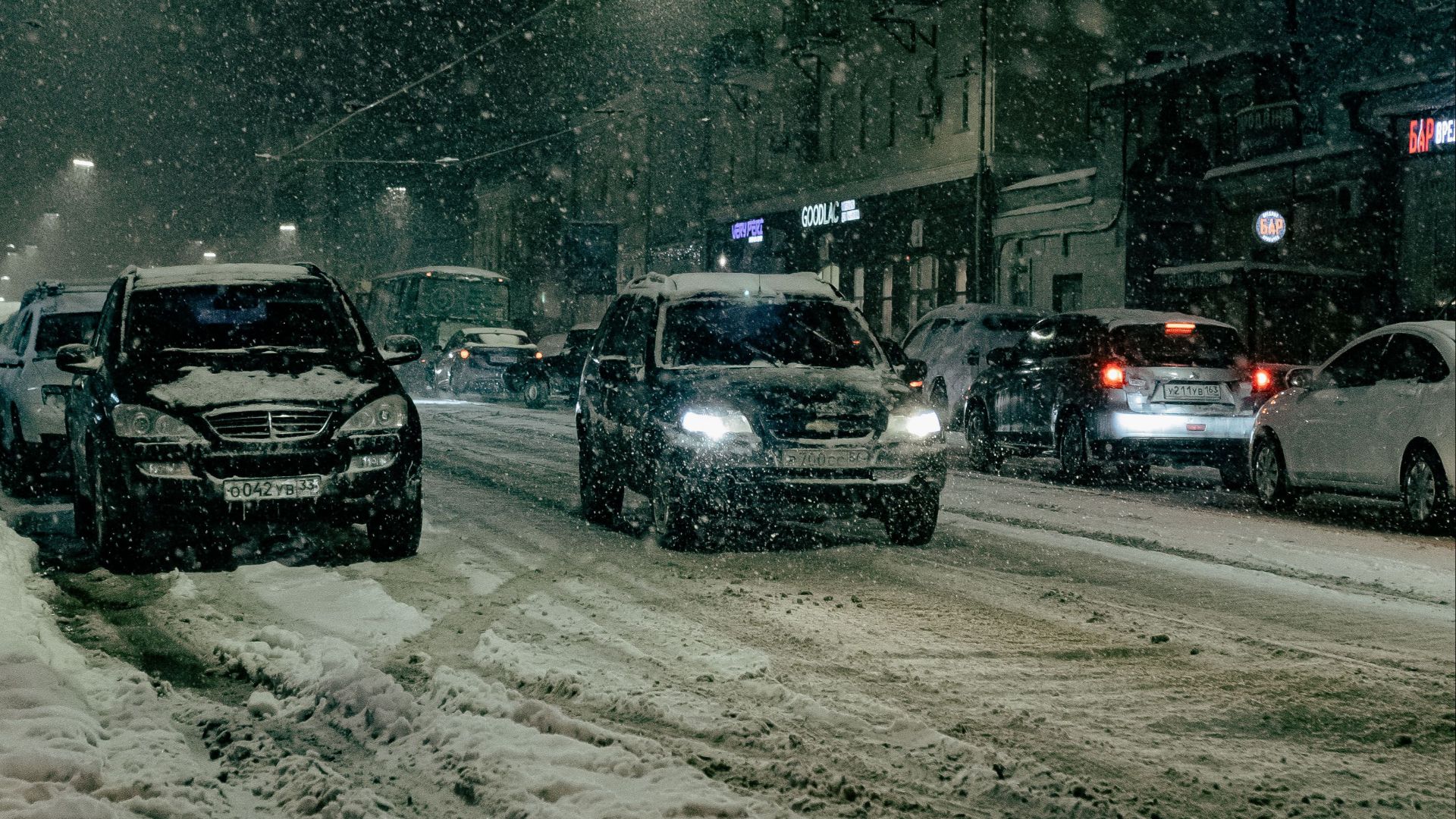 Cars driving on a snow-covered city street at night, heavy snowfall creates a moody atmosphere.