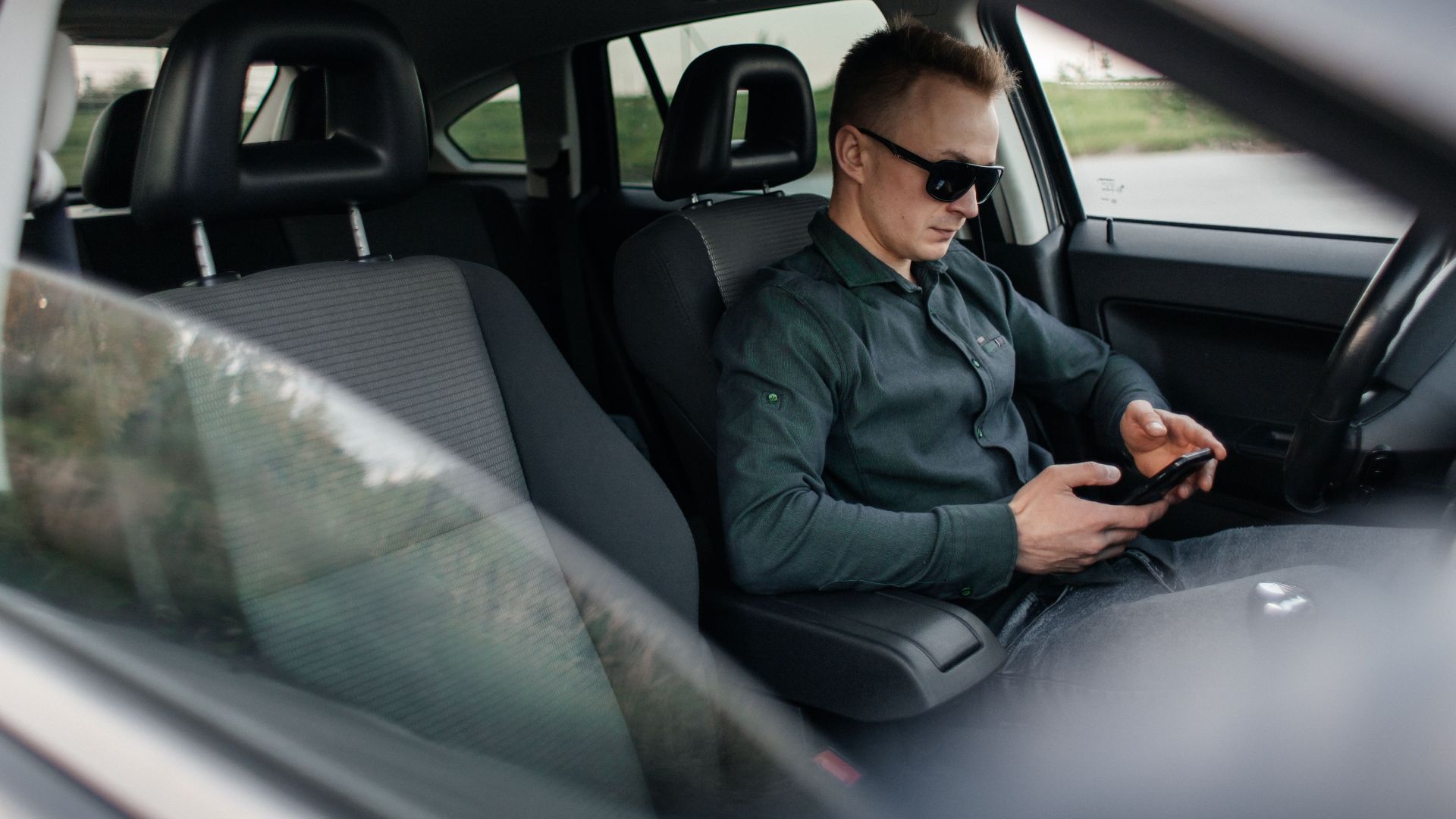 Young man using smartphone inside a parked car, wearing sunglasses.