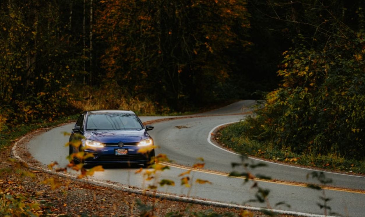 A blue car navigates a winding road surrounded by vibrant autumn foliage.