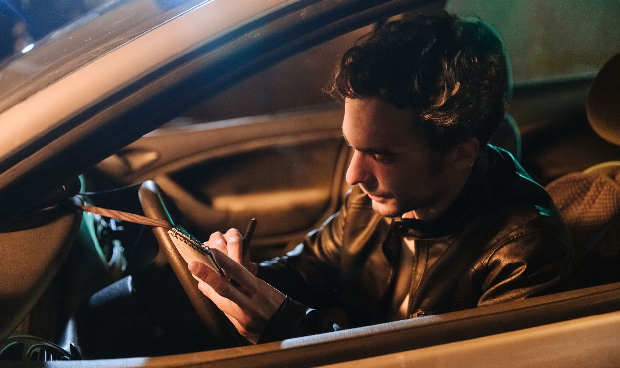 A man sitting in a car at night, writing notes in a notebook