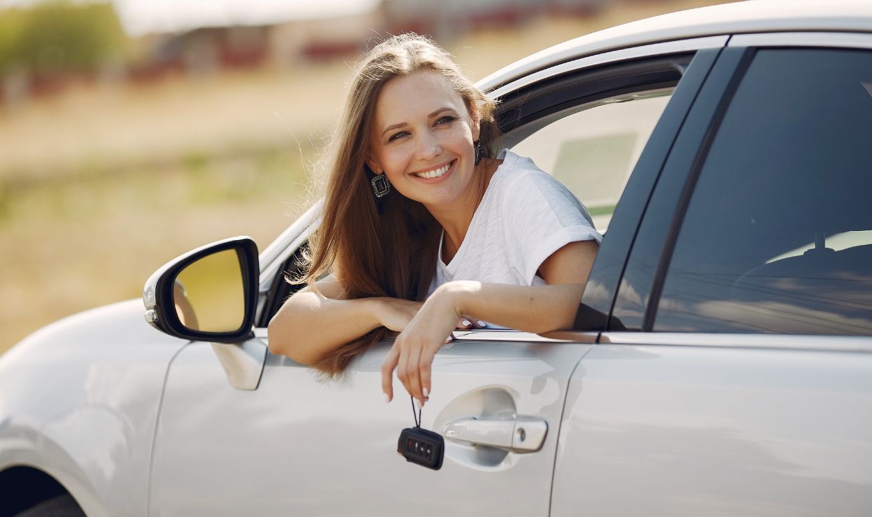 Female driver smiles while leaning out of her car window on a sunny day, embracing the joy of travel