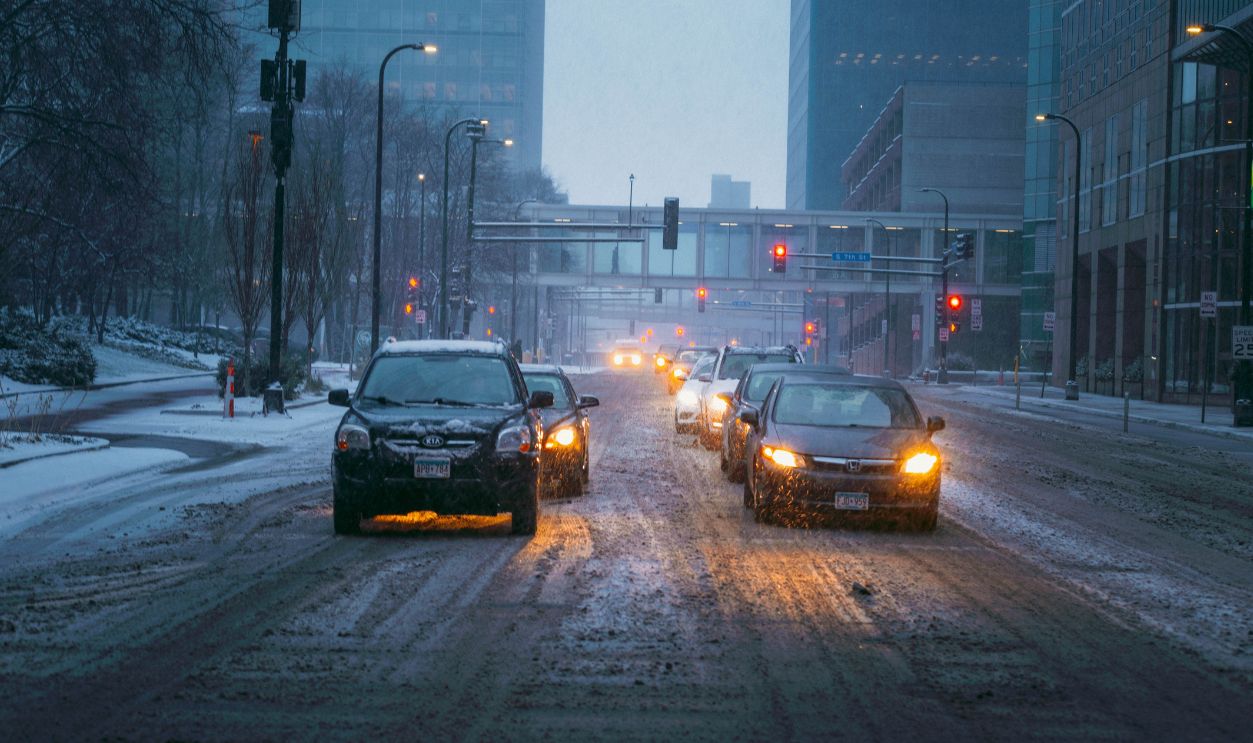 Cars driving on a snow-covered city street during an evening snowfall, with buildings lining the street