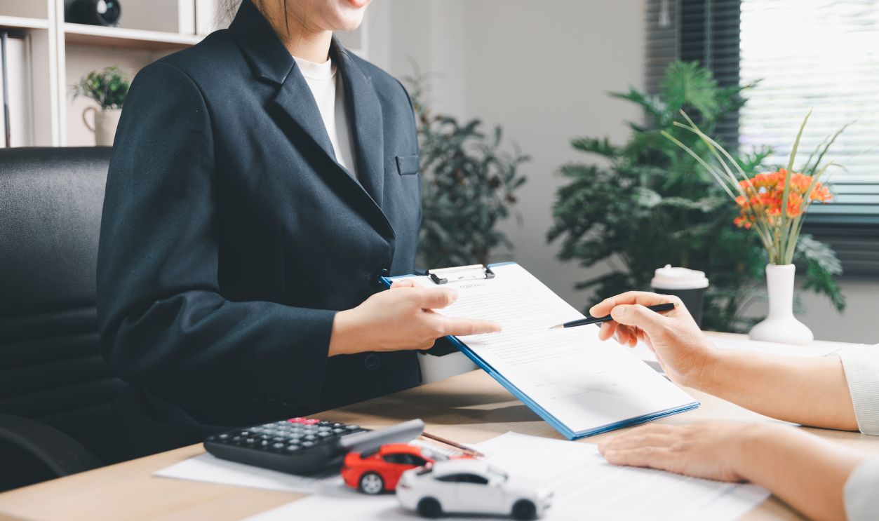 Smiling agent explaining auto loan contract to client at office desk with calculator and model cars, representing vehicle purchase consultation, payment planning, insurance options and trust.