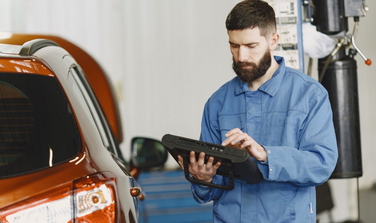 A mechanic in coveralls using a diagnostic tool at an auto repair shop beside a car.